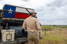 A man in a khaki outfit stands by a vehicle adorned with a Texas flag, holding a shotgun. An open cooler and a beer bottle are visible in the truck's bed. The background features a cloudy sky and grassland, while he proudly sports the Drewski Belt from Zilker Belts, crafted from world-class Argentine leather.