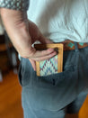 A person is placing the Drewski Wallet by Zilker Belts into the back pocket of their gray pants. The handcrafted leather wallet features a colorful woven design, and the person is wearing a patterned belt.