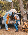 Two people in aprons lean over to check a large outdoor smoker on a trailer, their focus as meticulous as the hand-stitched details of Zilker Belts' Rambler piece. Nearby, a stack of firewood waits beneath the trees.